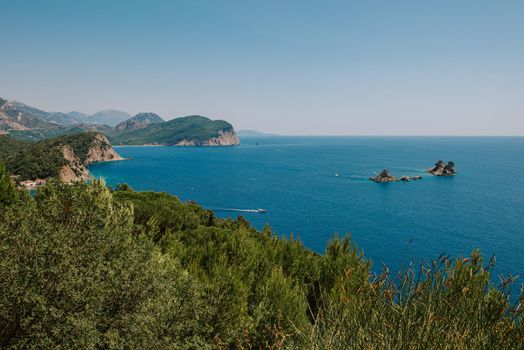 Picturesque view on rocks on a sunny day from the sea. Budva riviera, Montenegro. Aerial view of Sveti Nikola, Budva island, Montenegro. Hawaii beach, umbrellas and bathers and crystal clear waters. Jagged coasts with sheer cliffs overlooking the transparent sea. Wild nature. The rocky shore of Island St.Nikola near Budva, Montenegro. Picturesque summer view of Adriatic sea coast in Budva Riviera.