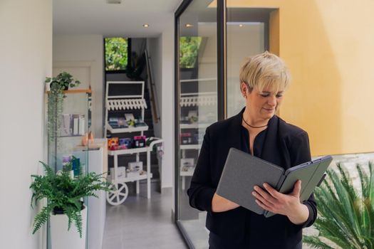 Smiling mature professional beautician with blonde hair, dressed in black work uniform, checking email on her Tablet. owner in her small beauty salon looking at laptop. Work from home, enterprising woman, active small business owner. Relaxed atmosphere and soft lighting from window, natural light, table, computer. Horizontal