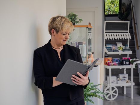 Smiling mature professional beautician woman with blonde hair, dressed in black work uniform, consulting visitors on her Tablet. Beautician in her small beauty salon looking at her laptop. Work from home, freelance, business, creative occupation, lifestyle concept. Relaxed atmosphere and soft lighting from window, natural light, table, computer. Horizontal.