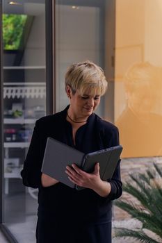 Smiling mature professional beautician woman with blonde hair, dressed in black work uniform, jotting down client appointments on her tablet. worker in business hallway consulting her laptop. Work from home, freelance, business, creative occupation, lifestyle concept. Relaxed atmosphere and soft lighting from window, natural light, table, computer. Vertical