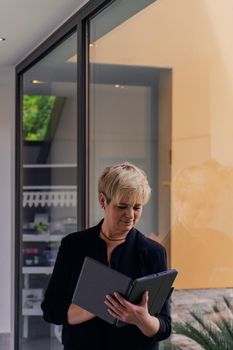 Smiling mature professional beautician woman with blonde hair, dressed in black work uniform, consulting visitors on her Tablet. Beautician in her small beauty salon looking at her laptop. Work from home, freelance, business, creative occupation, lifestyle concept. Relaxed atmosphere and soft lighting from window, natural light, table, computer. Vertical