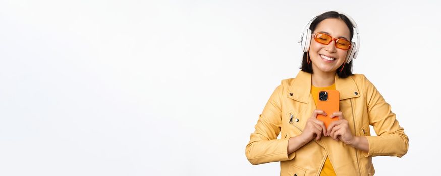 Technology concept. Stylish asian girl in headphones, holding smartphone, dancing and singing, listening music, standing over white background.