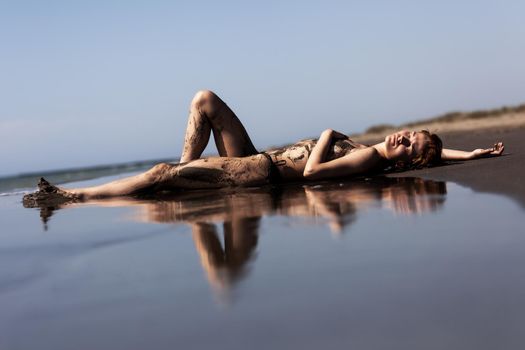 Sexy female body covered with sand. Girl on a beach with sea water reflection.