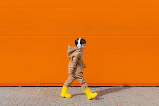 A boy listens to music with headphones on the background of an orange wall . Copy space. Mockup. An article about modern children. An article about modern music. Article about the choice of headphones. Urban lifestyle