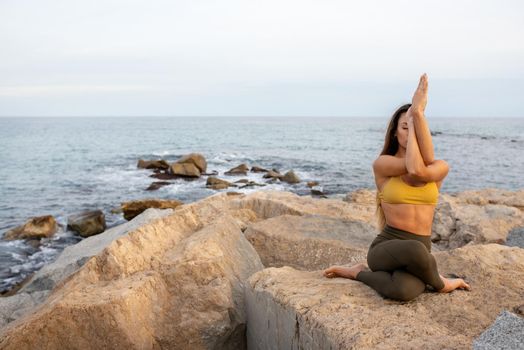 Young woman doing gomukhasana with garudasana arms. Yoga practice in nature near the ocean.Copy space. Healthy lifestyle and spirituality concept.