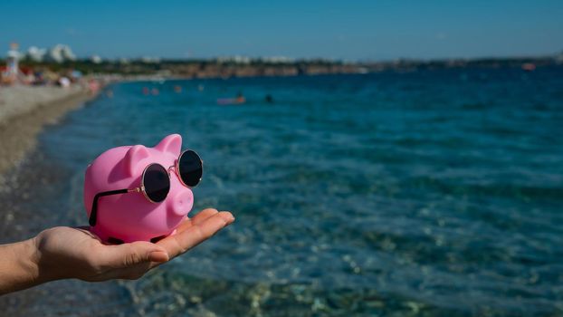 A woman holds a piggy bank in sunglasses on a pebble beach near the sea. Budget vacation