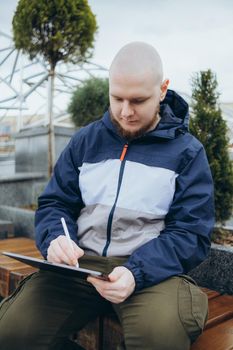 From below of hipster man using modern tablet with stylus while creating new project and sitting on street near glass skyscraper