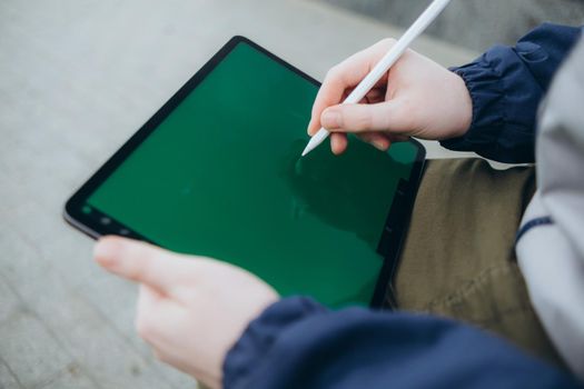 From below of hipster man using modern tablet with stylus while creating new project and sitting on street near glass skyscraper