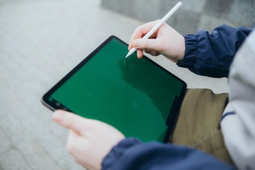 From below of hipster man using modern tablet with stylus while creating new project and sitting on street near glass skyscraper