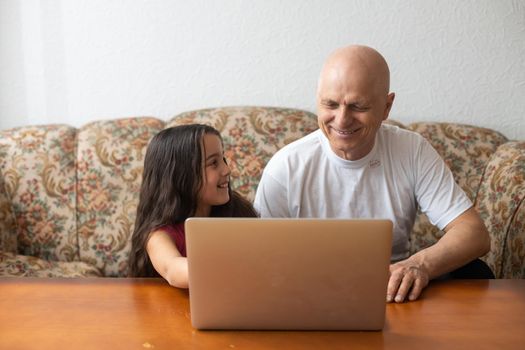 Adorable little girl hugging happy grandfather using laptop at home