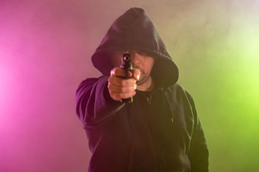 Man in black hoodie points handgun in colored haze of smoke