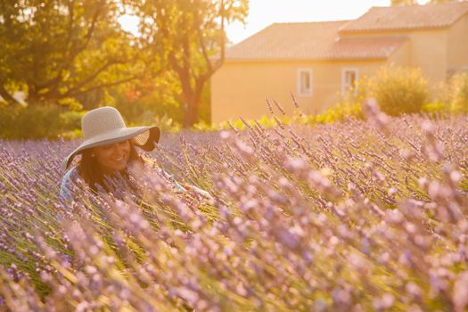 The beautiful young girl in a blue dress and cap sits in a lavender flowers, long curly hair, smile, pleasure, a house of the gardener in the background, trees, perspective of a lavender. High quality photo