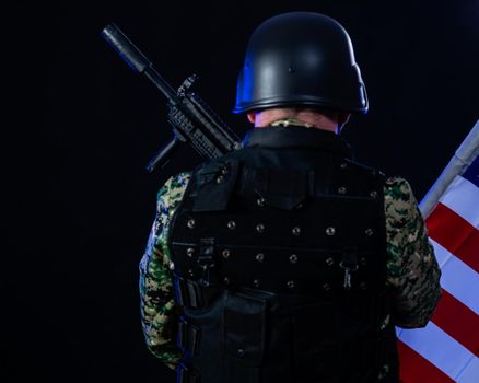 Soldier in army fatigues holding assault rifle and US flag, from back