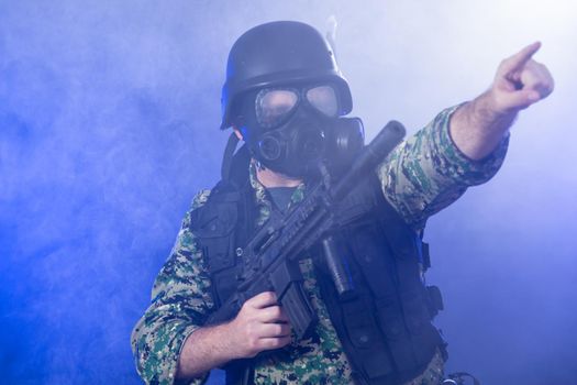 Soldier in army fatigues wearing gas mask holding assault rifle, points through haze of blue smoke