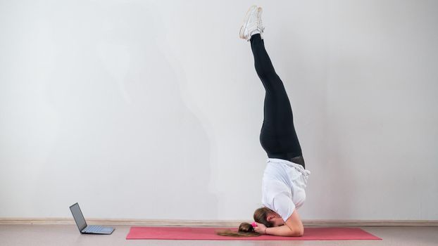 A chubby young woman watches an online yoga lesson on a laptop. Distance sports training. Forearm stand.