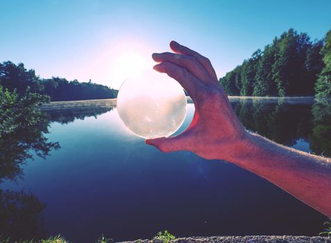 Looking through glass lens. Hand hold glass ball withuniquely reflects  summer lake scene with sunset.