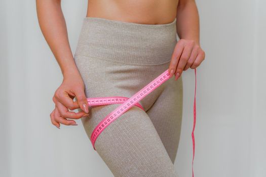 Cropped view of slim woman measuring her leg with tape measure at home, close-up. An unrecognizable European woman checks the result of a weight loss diet or liposuction indoors. Healthy lifestyle