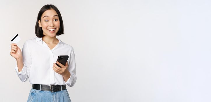 Online shopping concept. Image of young asian modern woman holding credit card and smartphone, buying with smartphone app, paying contactless, standing over white background.