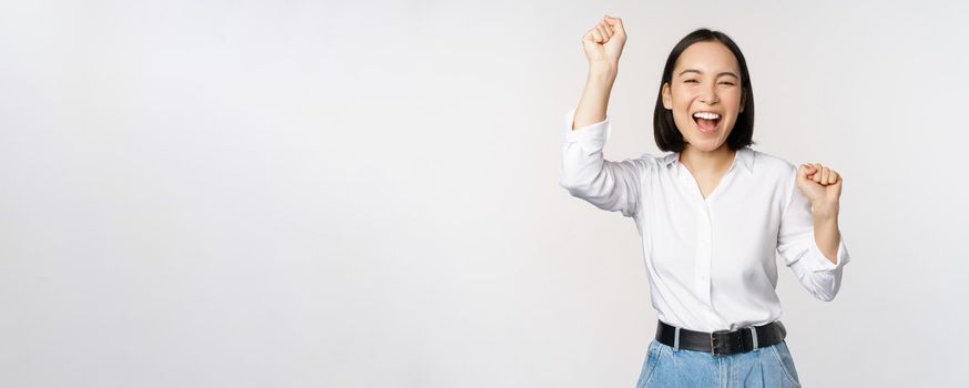 Image of happy lucky asian woman hooray gesture, winning and celebrating, triumphing, raising hadns up and laughing, standing over white background.