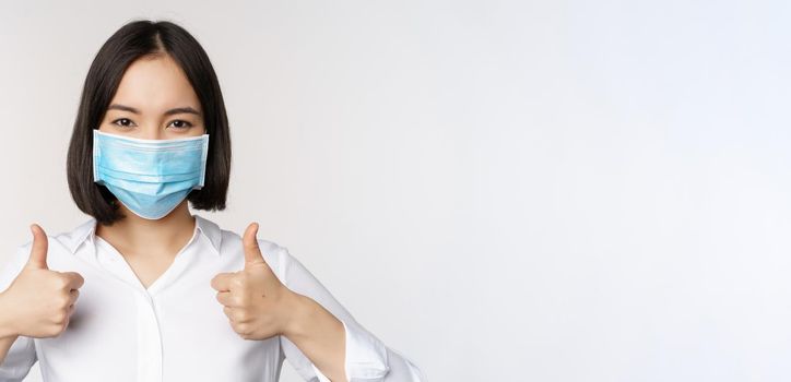 Portrait of smiling asian office lady in medical face mask, showing thumbs up, recommending smth, standing over white background.