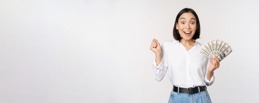 Enthusiastic young asian woman looking excited at camera, holding money dollars in hand, standing over white background.