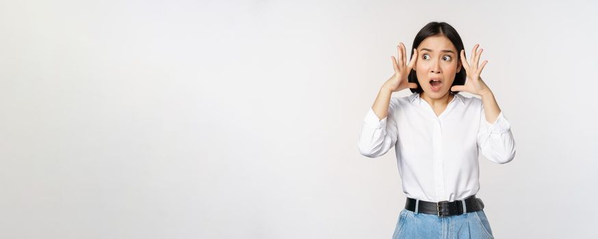 Image of shocked anxious asian woman in panic, holding hands on head and worrying, standing frustrated and scared against white background.