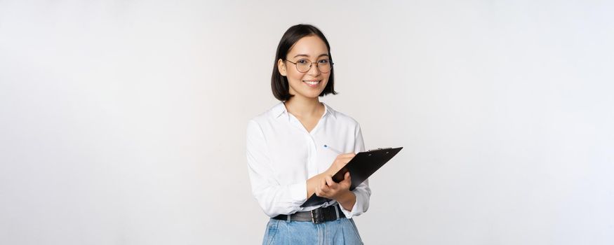 Image of smiling korean office lady, company employee writing down on clipboard, taking notes, standing over white background.