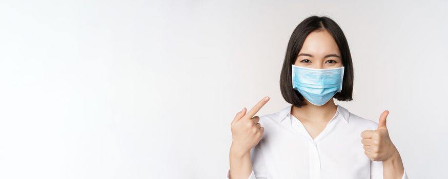 Portrait of asian girl in medical mask showing thumbs up sign and pointing at her covid protection, standing over white background.