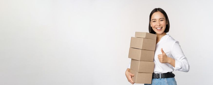 Image of happy modern asian woman showing thumbs up, holding boxes delivery goods, standing against white background.