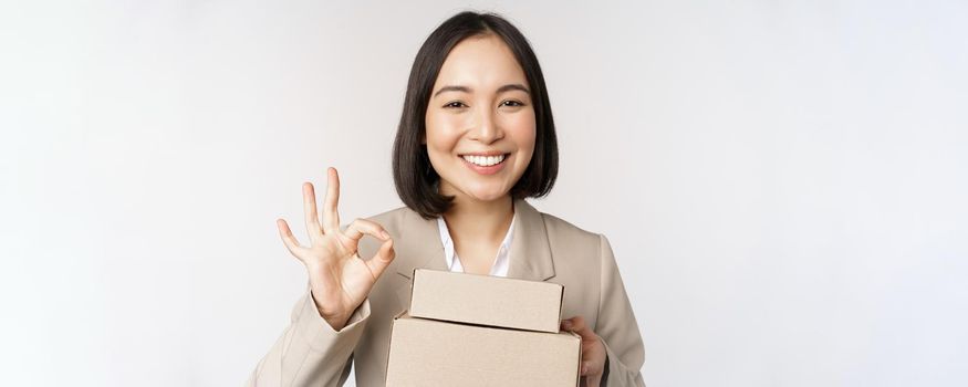 Smiling asian businesswoman, showing okay sign and boxes with delivery goods, prepare order for client, standing over white background.