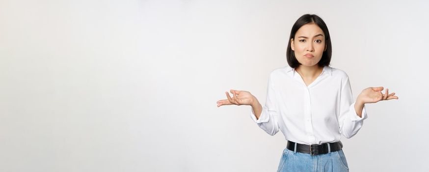 Image of confused asian office manager, office lady shrugging shoulders and looking clueless, standing puzzled against white background.