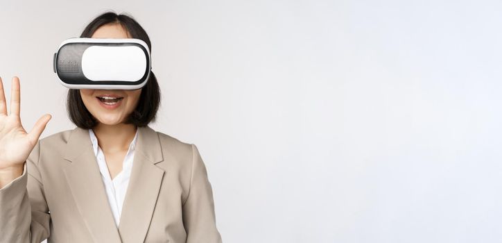 Meeting in vr chat. Asian businesswoman in virtual reality glasses, waving hand and saying hello, greeting someone, standing over white background.