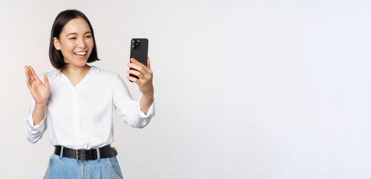 Image of stylish modern asian woman waving hand at smartphone front camera, video chat, chatting with person on mobile phone application, white background.