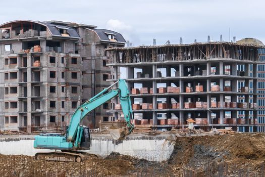 Excavator with bucket works on construction site against background of houses under construction