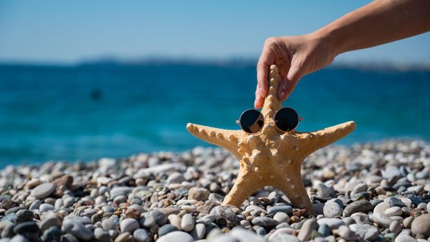 Woman holding a starfish in sunglasses on a pebble beach by the sea