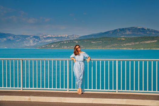 The beautiful girl in a blue dress and sunglasses poses on the bridge, the long chestnut hair, happy and smiles, azure water of the lake and slopes of mountains on a background. High quality photo