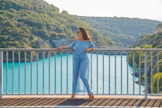 The beautiful girl in a blue dress and sunglasses poses on the bridge, the long chestnut hair, happy and smiles, azure water of the lake and slopes of mountains on a background. High quality photo