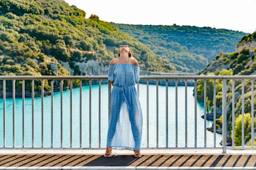 The beautiful girl in a blue dress and sunglasses poses on the bridge, the long chestnut hair, happy and smiles, azure water of the lake and slopes of mountains on a background. High quality photo