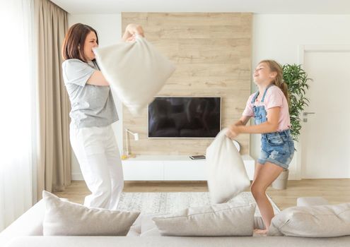 Happy family games. Single mother and her child girl are fighting pillows and jumping on a couch
