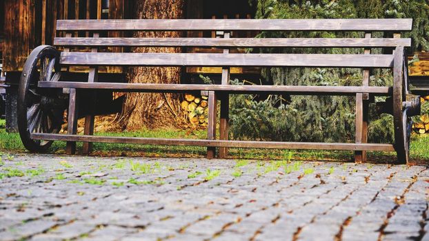 a long seat for several people, typically made of wood or stone.Wooden vintage bench with cart wheels on paving stones.