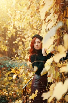 Woman in knitted sweater among yellow fall leaves. Portrait of young red-haired girl with beret in autumn park. Warm sunny weather. Stylish model in nature.