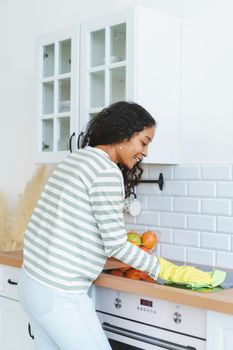 Smiling African-American woman occupied with household duties in kitchen. Sweeping dust and removing dirt in gloves with washcloth from modern light kitchenette unit. Vertical, copy space