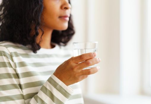 Cropped African-American woman holding glass of clear water and looking at window. Healthy and refreshing start of day. Natural daylight. Soft focused background