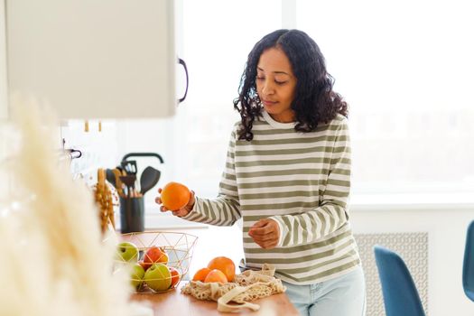 African-American woman sorting out fruit in kitchen. Choosing healthy vegetarian food. Vegan lifestyle. Looking at ripe oranges and apples. Taking out of eco-bag. Natural daylight, horizontal