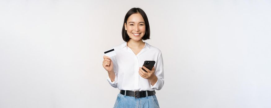 Online shopping concept. Image of young asian modern woman holding credit card and smartphone, buying with smartphone app, paying contactless, standing over white background.