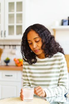 Tired and sick young African-American female dealing with terrible pain. Concept of taking fizz pill to cure disease. Looking at glass of water while sitting in kitchen. Vertical