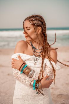 Model in boho style in a white long dress and silver jewelry on the beach. Her hair is braided, and there are many bracelets on her arms