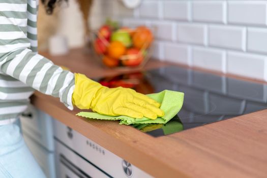 Anonymous woman wearing yellow rubber gloves leaning stove with green wathcloth. Concept of doing household chores and removing dirt from kitchen surface. Disinfecting and sanitizing