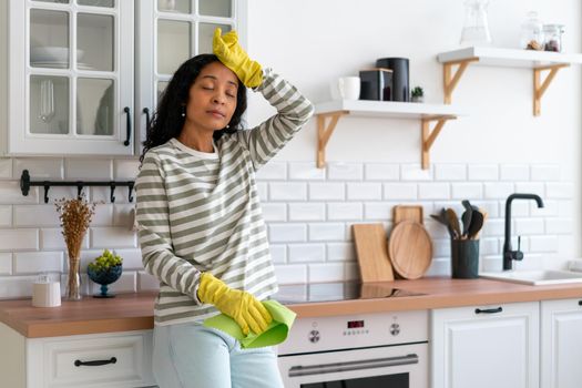 African-american woman wiping sweat from forehead after cleaning kitchen unit. Having rest after dust handling in rubber gloves with washcloth. Concept of tired housekeeper. Finishing cleanup