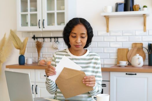 African-american woman writing and signing postcard in kitchen. Preparing correspondence for mail delivery. Concept of sending letters. Holiday invations. Searching for ideas online on laptop